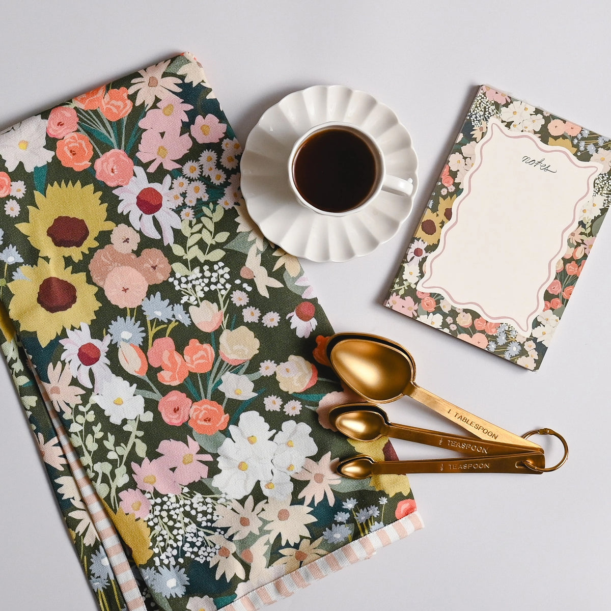 A styled flat-lay featuring a floral kitchen towel, a cup of black coffee on a white saucer, a floral notepad, and gold measuring spoons on a light gray background.