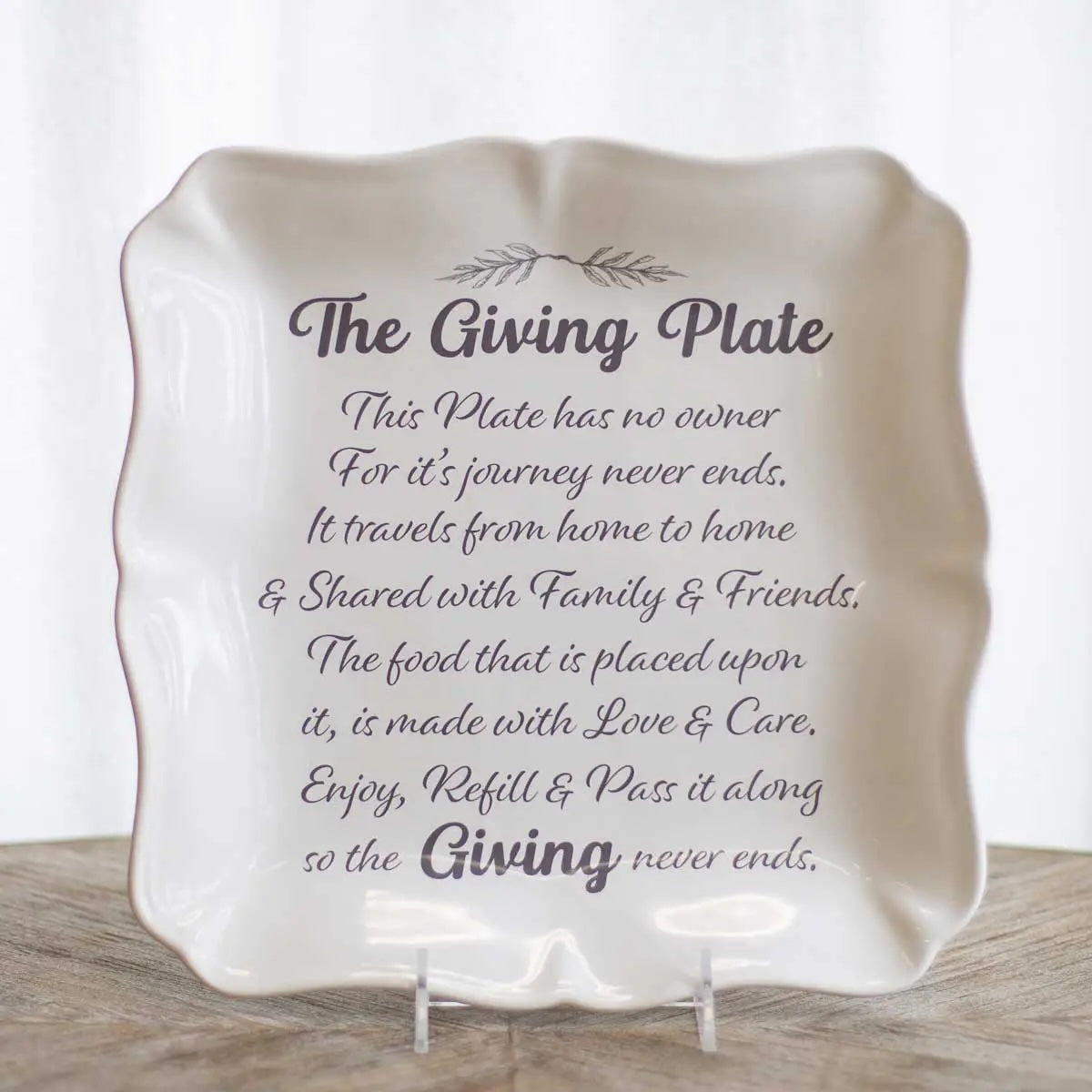A square ceramic giving plate with scalloped edges, displayed on small clear stands. The plate is off-white with dark gray printed text that reads 'The Giving Plate' followed by a sentimental message about sharing and giving.