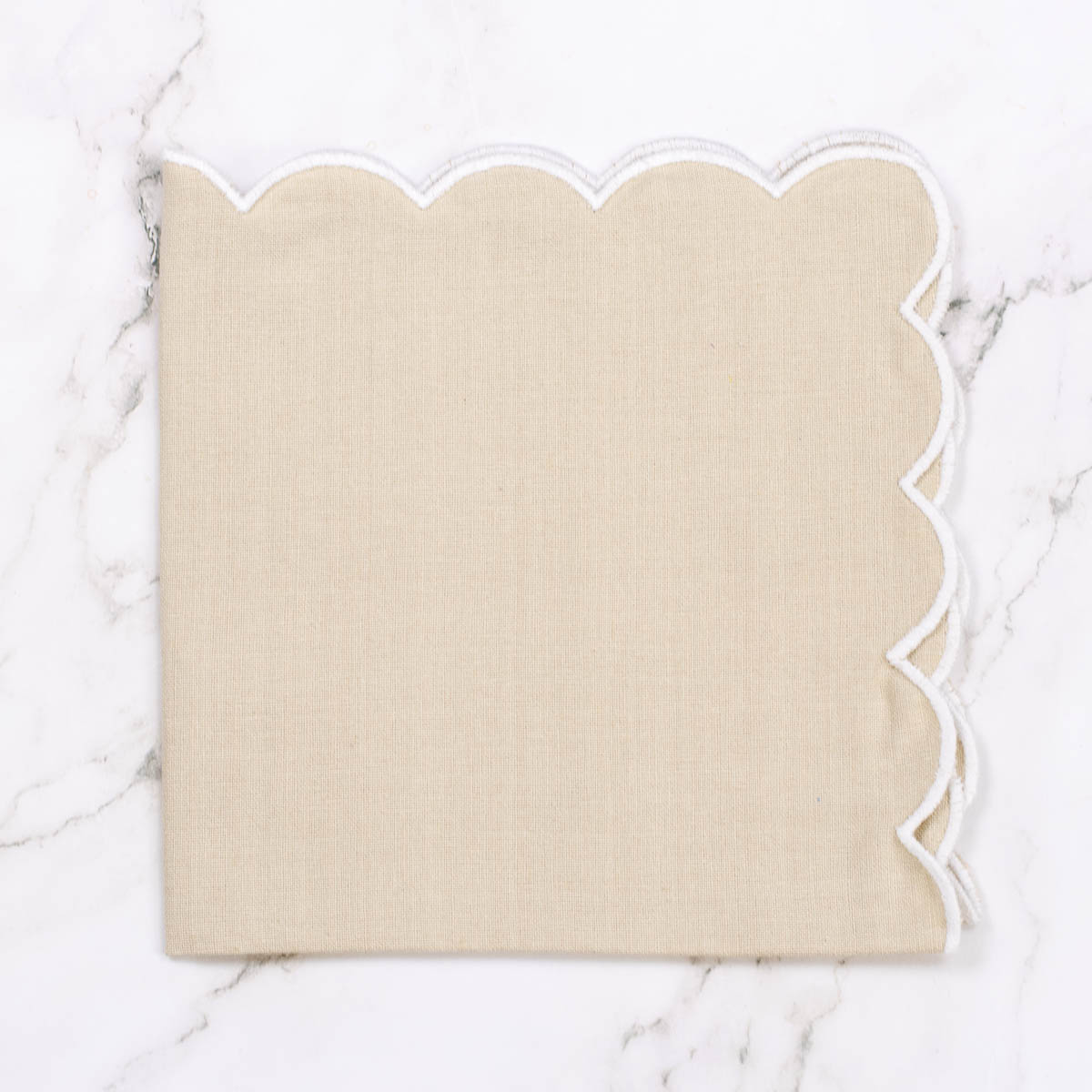 A square beige napkin with a scalloped white edge, laid flat on a white marble surface.