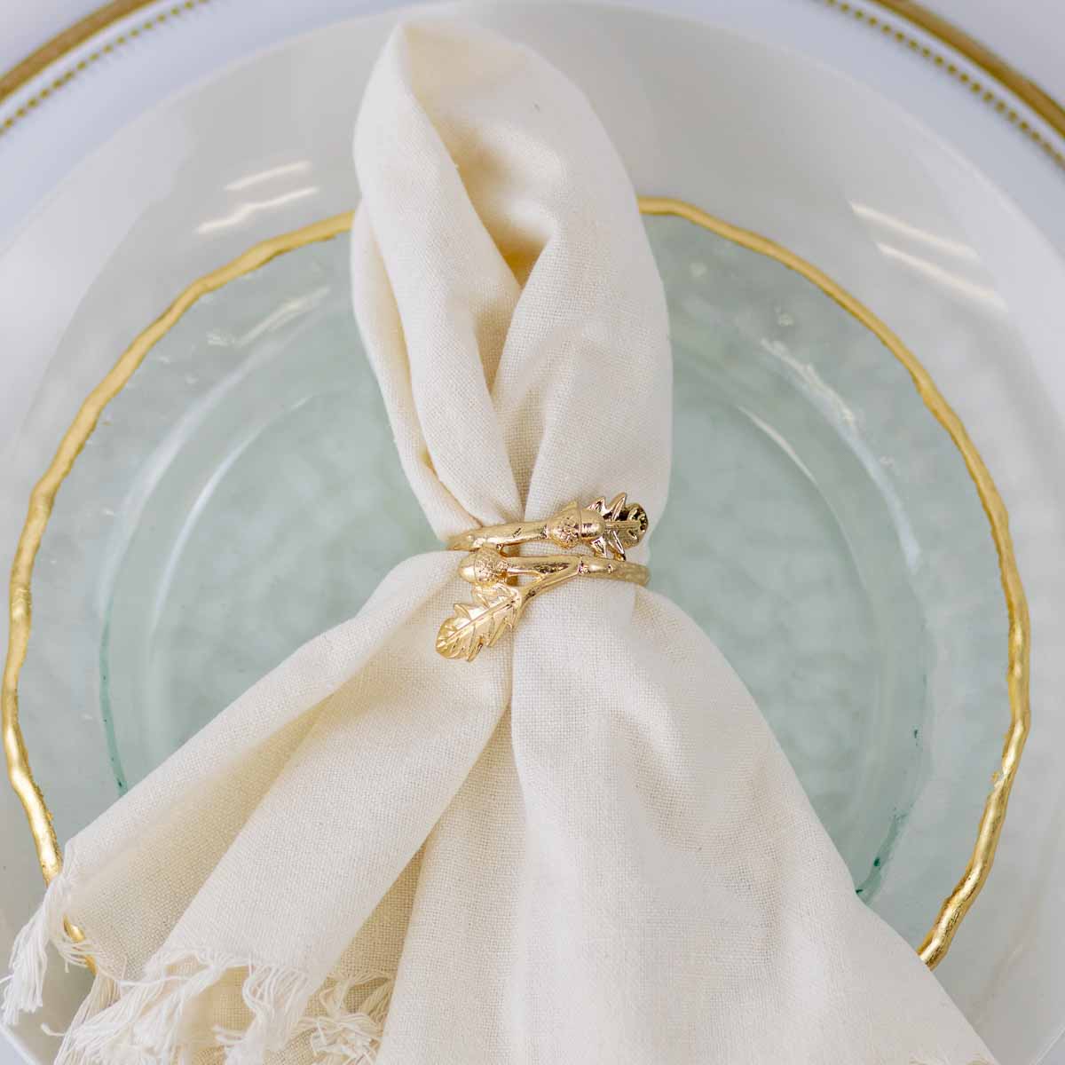 A cream-colored linen napkin tied with a gold leaf-shaped ring, resting on a clear glass plate with a gold-trimmed white ceramic plate.