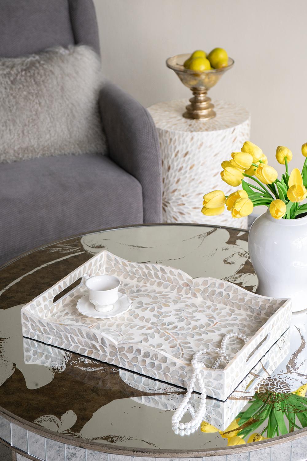 Round glass coffee table with decorative tray, white vase with yellow tulips, and fruit bowl in a living room setting.