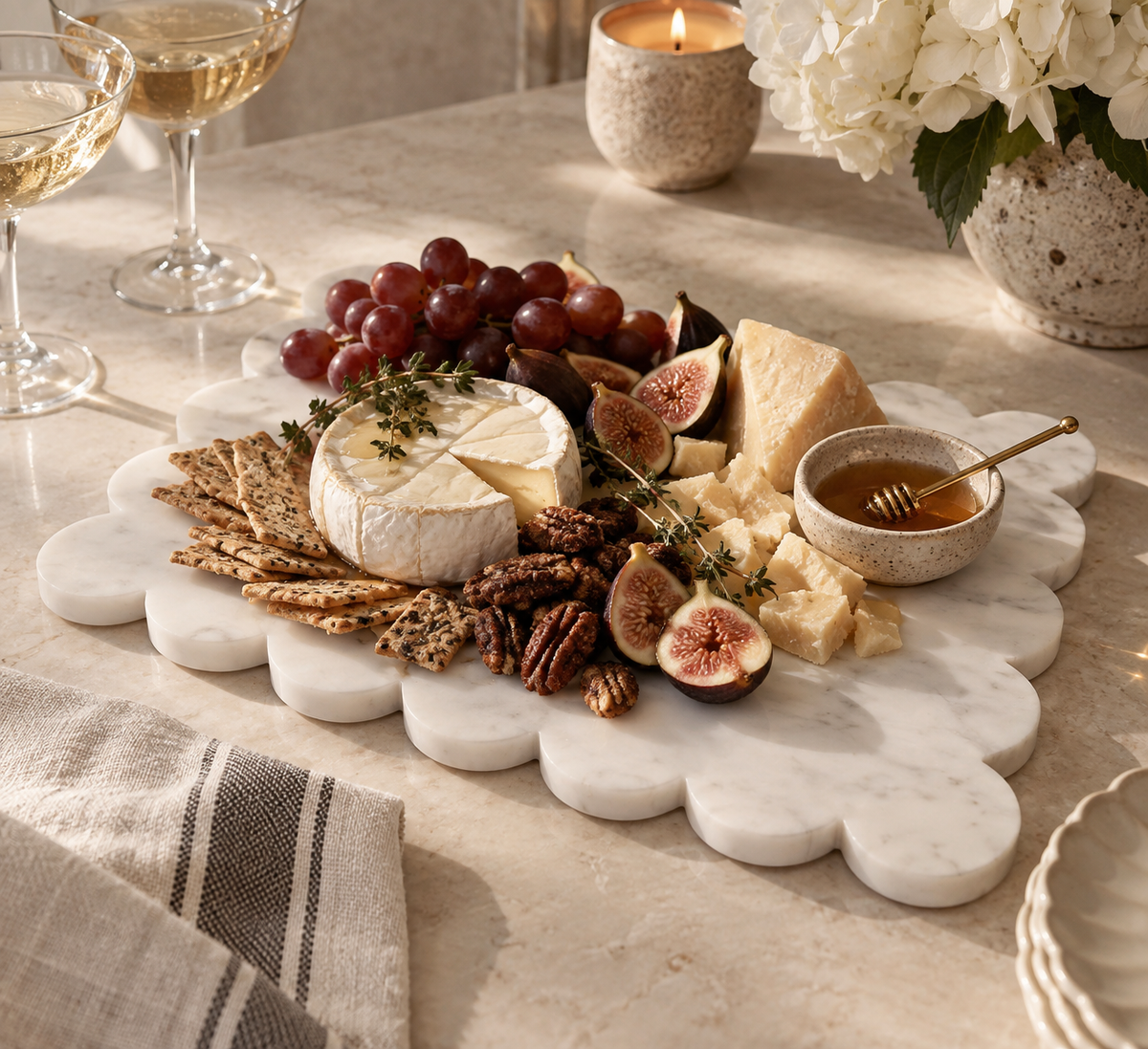 A scalloped white marble charcuterie board styled with cheese, grapes, figs, crackers, nuts, and a small honey bowl on a dining table, with wine glasses, a candle, and flowers in the background.