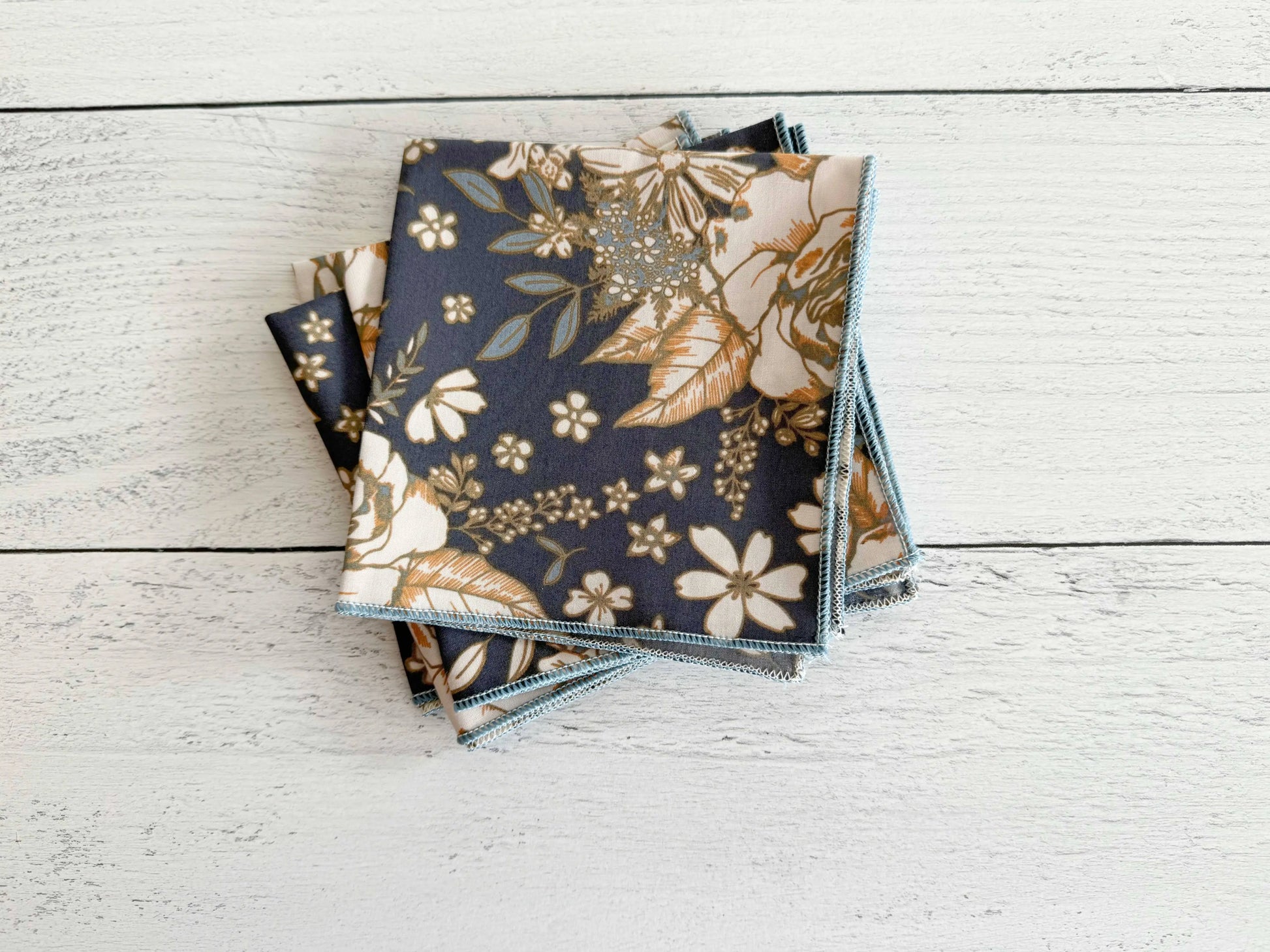 Set of navy fabric napkins with gold floral pattern and blue stitched edges, folded stack