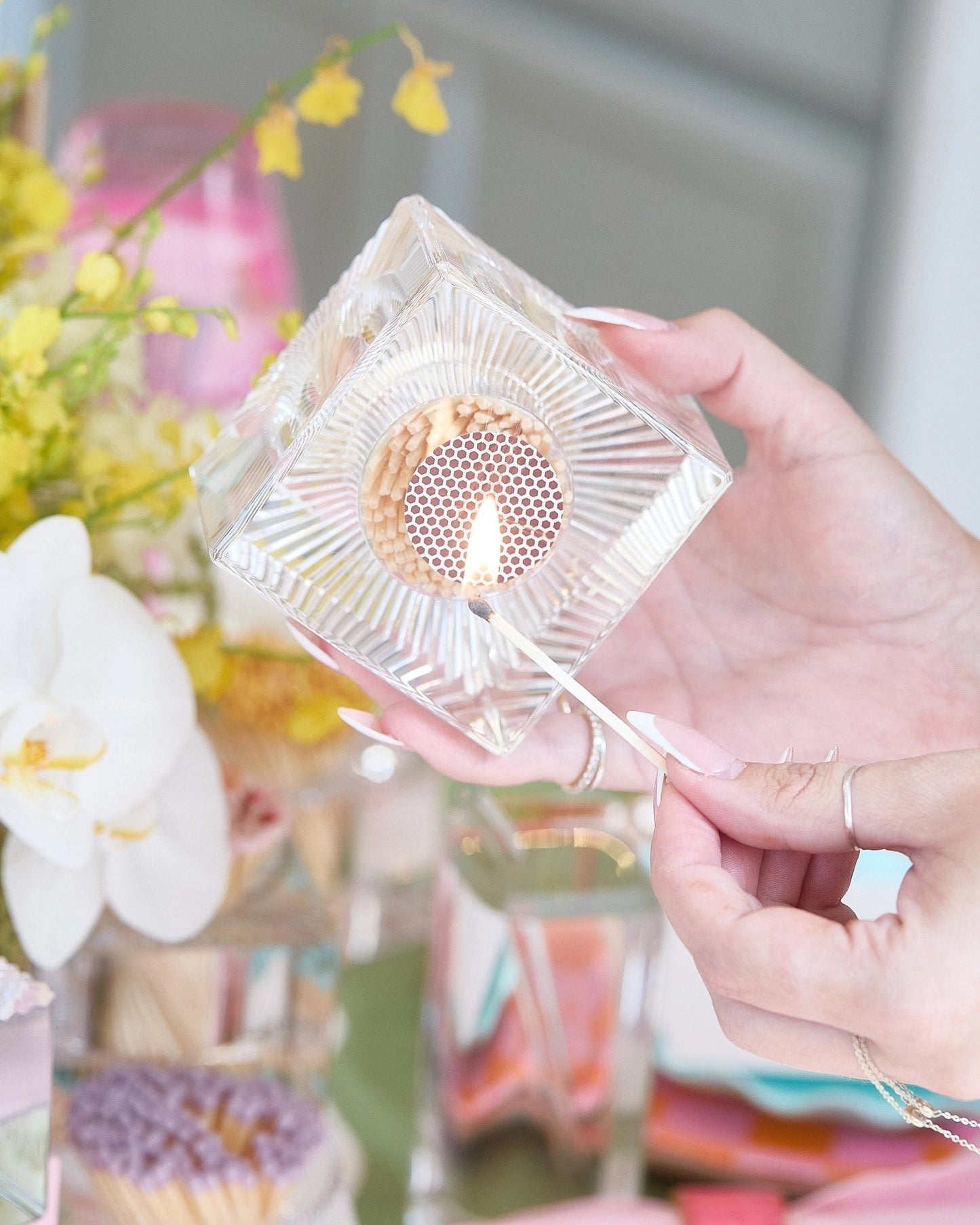 Clear acrylic match holder with striker, a lid held by a hand against a blurred floral background.