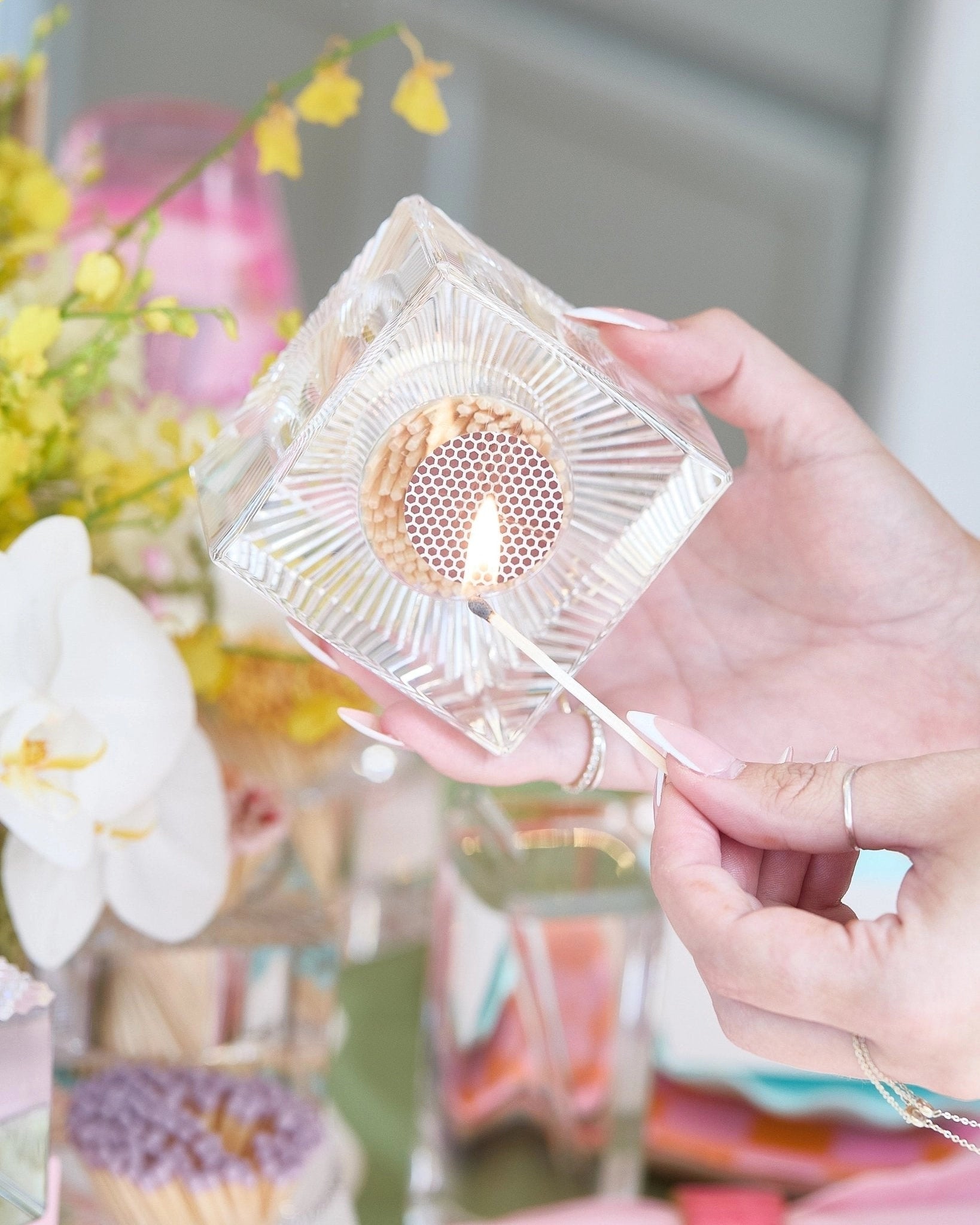 Clear acrylic match holder with striker, a lid held by a hand against a blurred floral background.