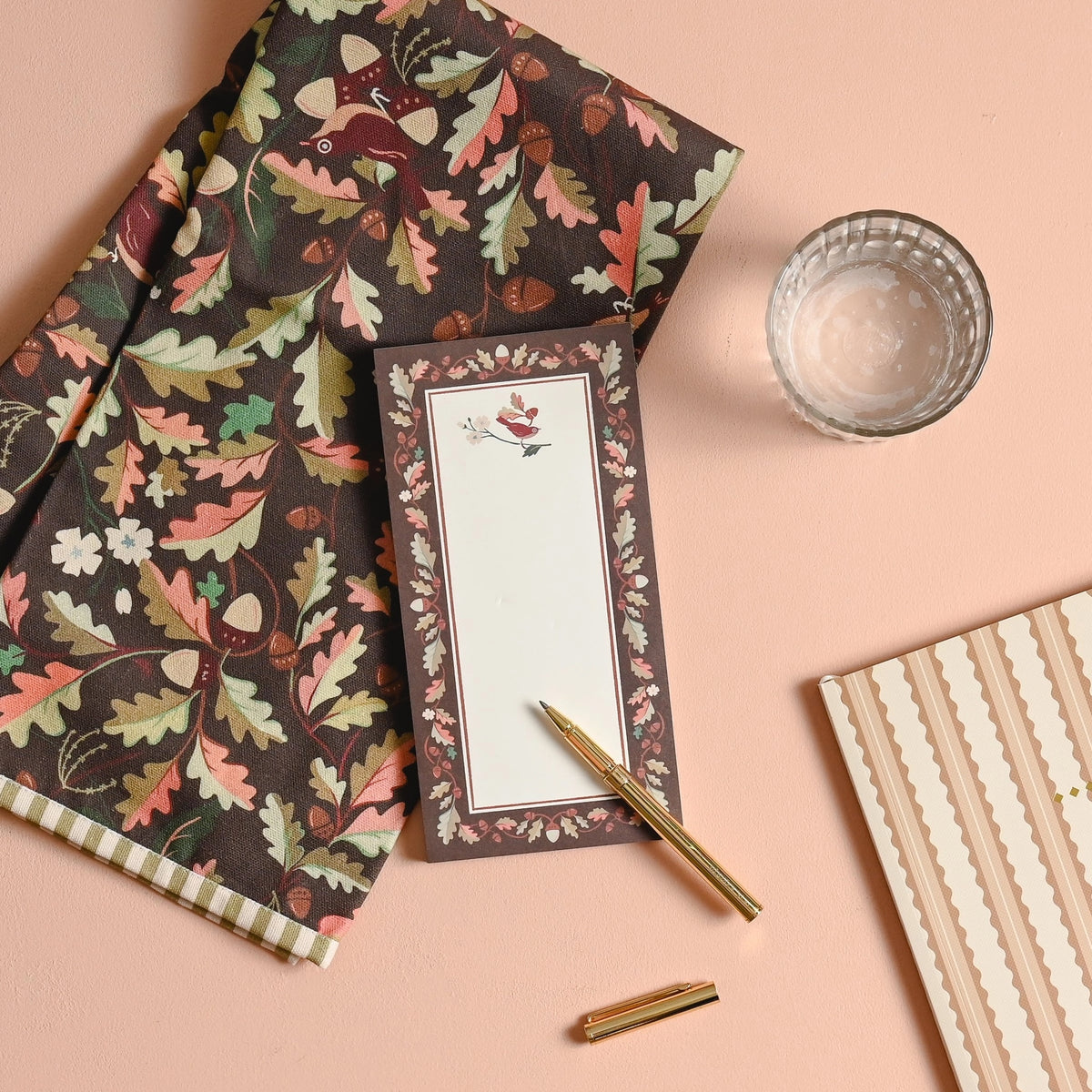 A styled flat-lay featuring a dark brown fabric napkin or tablecloth with a detailed autumn leaf and acorn pattern, alongside a matching floral notepad, gold pens, a glass of water, and a striped notebook on a solid peach background.