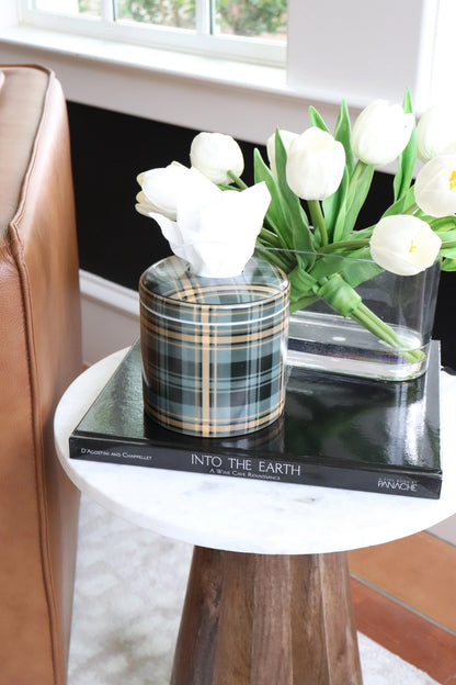 A tissue box cover in a green and black plaid pattern sits on a black book on a small round white marble side table, next to a glass vase of white tulips.