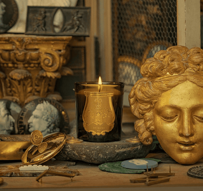 Trudon Cyrnos Candle with a decorative lid displayed in an ornate setting with gold artifacts and a stone surface.