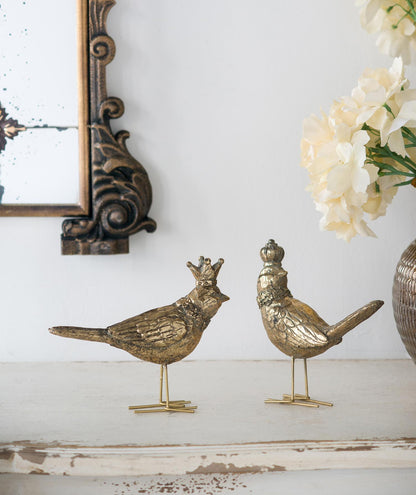 Two decorative brass birds on a white surface with a mirror and flowers in the background.