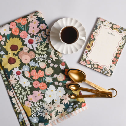 A styled flat-lay featuring a floral kitchen towel, a cup of black coffee on a white saucer, a floral notepad, and gold measuring spoons on a light gray background.