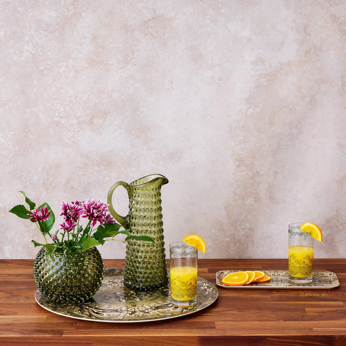 Green textured pitcher and glasses with orange juice on a wooden table against a beige wall.