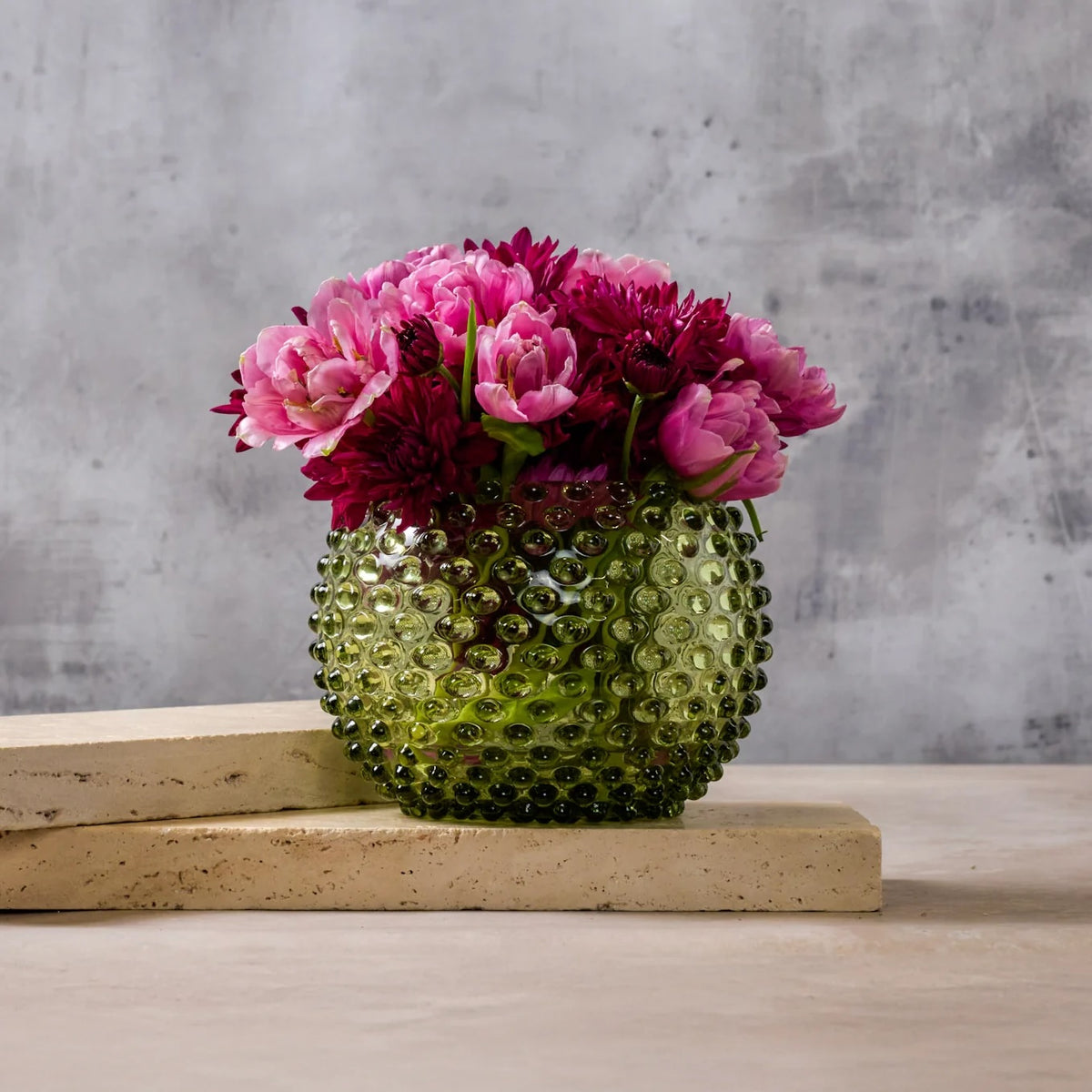 A textured green glass vase filled with pink and burgundy flowers, displayed on a light beige stone block against a gray concrete wall.