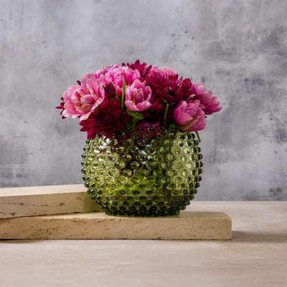 A textured green glass vase filled with pink and burgundy flowers, displayed on a light beige stone block against a gray concrete wall.