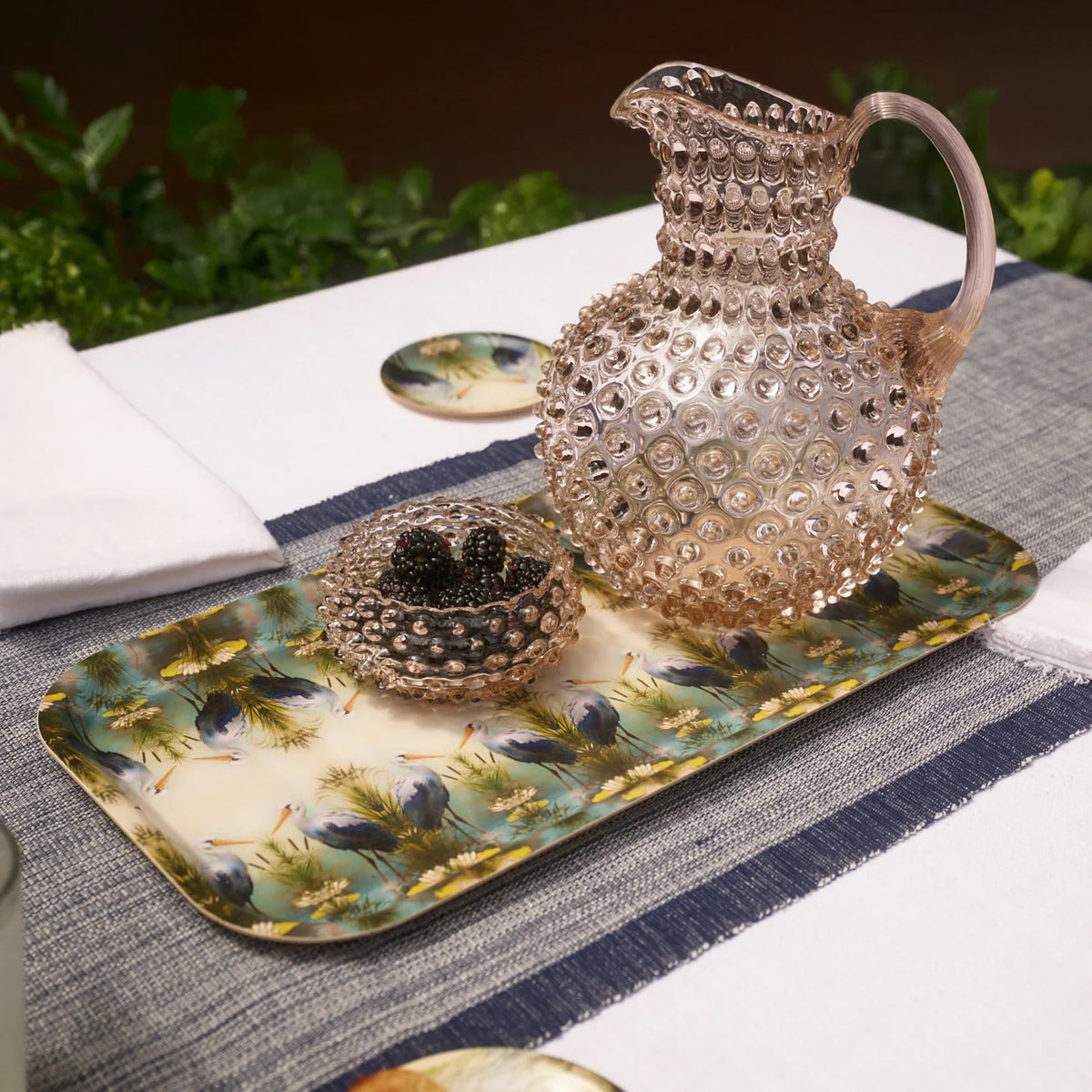 A decorative glass pitcher with a textured hobnail pattern sits on a matching patterned tray alongside a small glass bowl of blackberries on a dining table setting.
