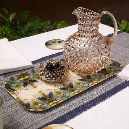 A decorative glass pitcher with a textured hobnail pattern sits on a matching patterned tray alongside a small glass bowl of blackberries on a dining table setting.