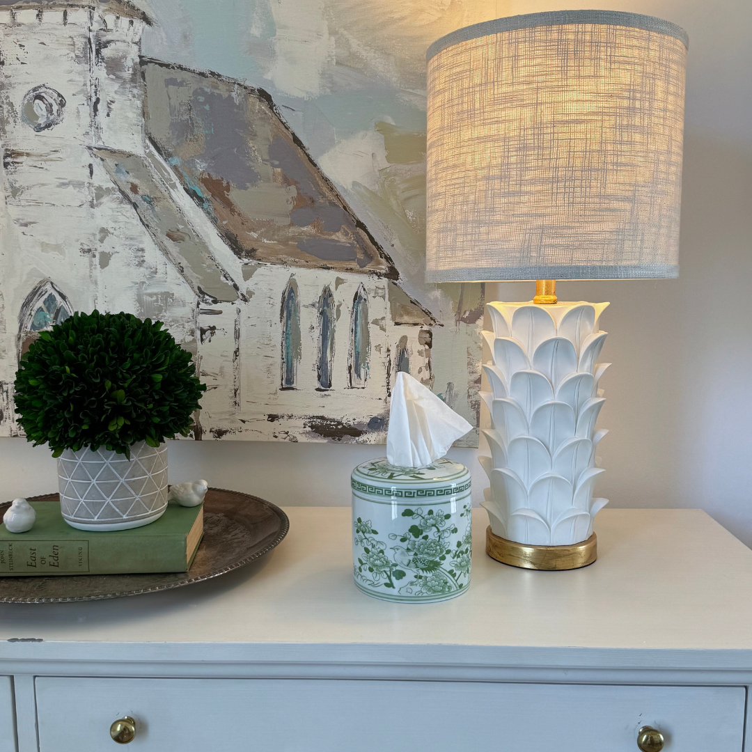 Decorative items on a dresser including a lamp, tissue box, and potted plant with a painting of a church in the background.