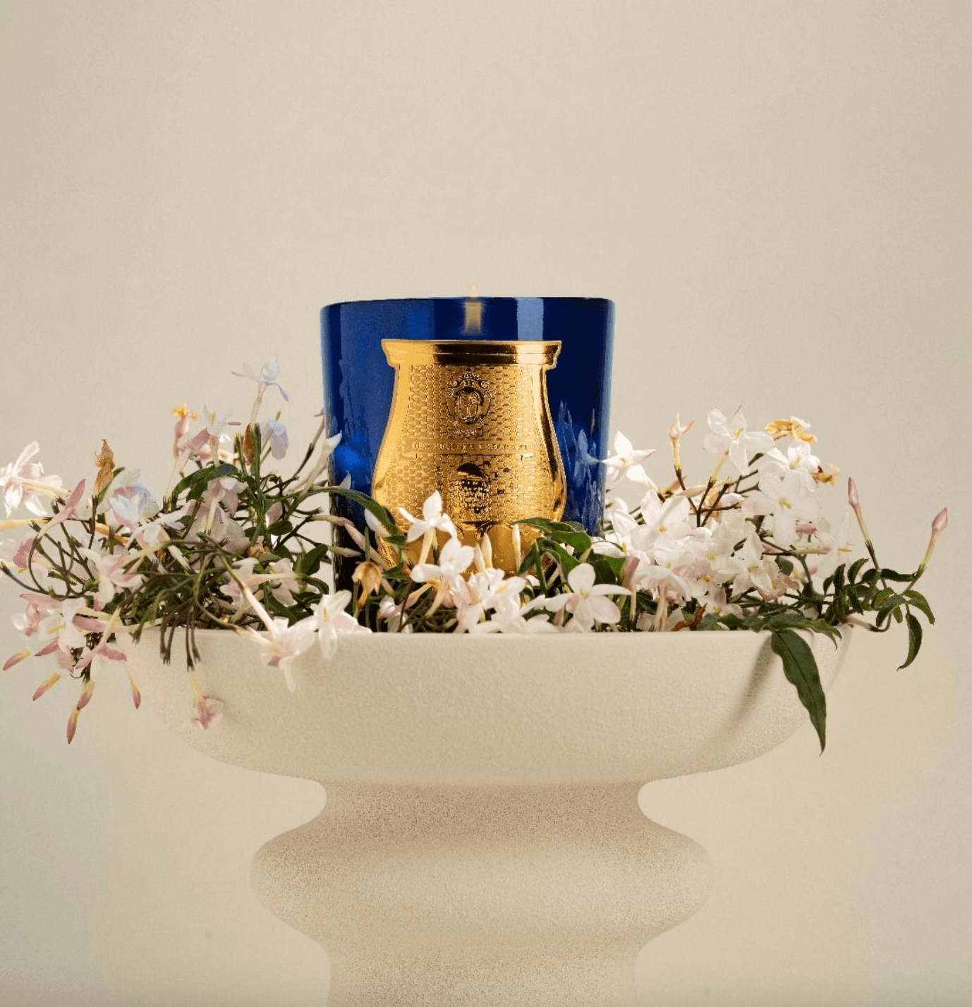 Trudon Madurai Candle in a blue and gold glass vessel styled in a white vase with flowers on a beige background.