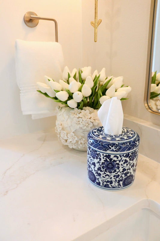 Bathroom counter with white tulips in a vase, blue and white ceramic tissue holder, and white towel.