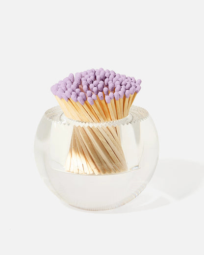 A clear round match holder container holding a bundle of wooden matchsticks with light purple tips, photographed on a clean white background.