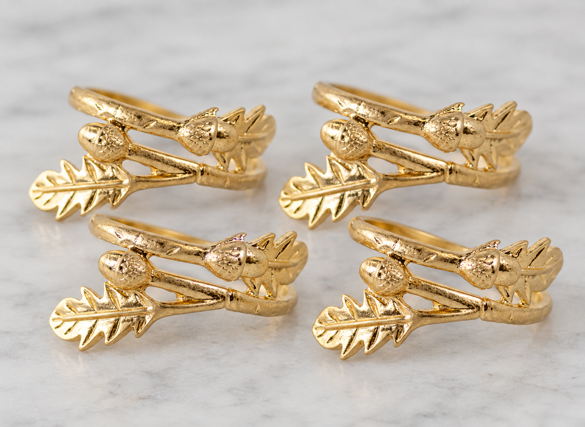 Four gold rings with a textured leaf and acorn design, arranged in a 2x2 grid on a light gray marble surface.