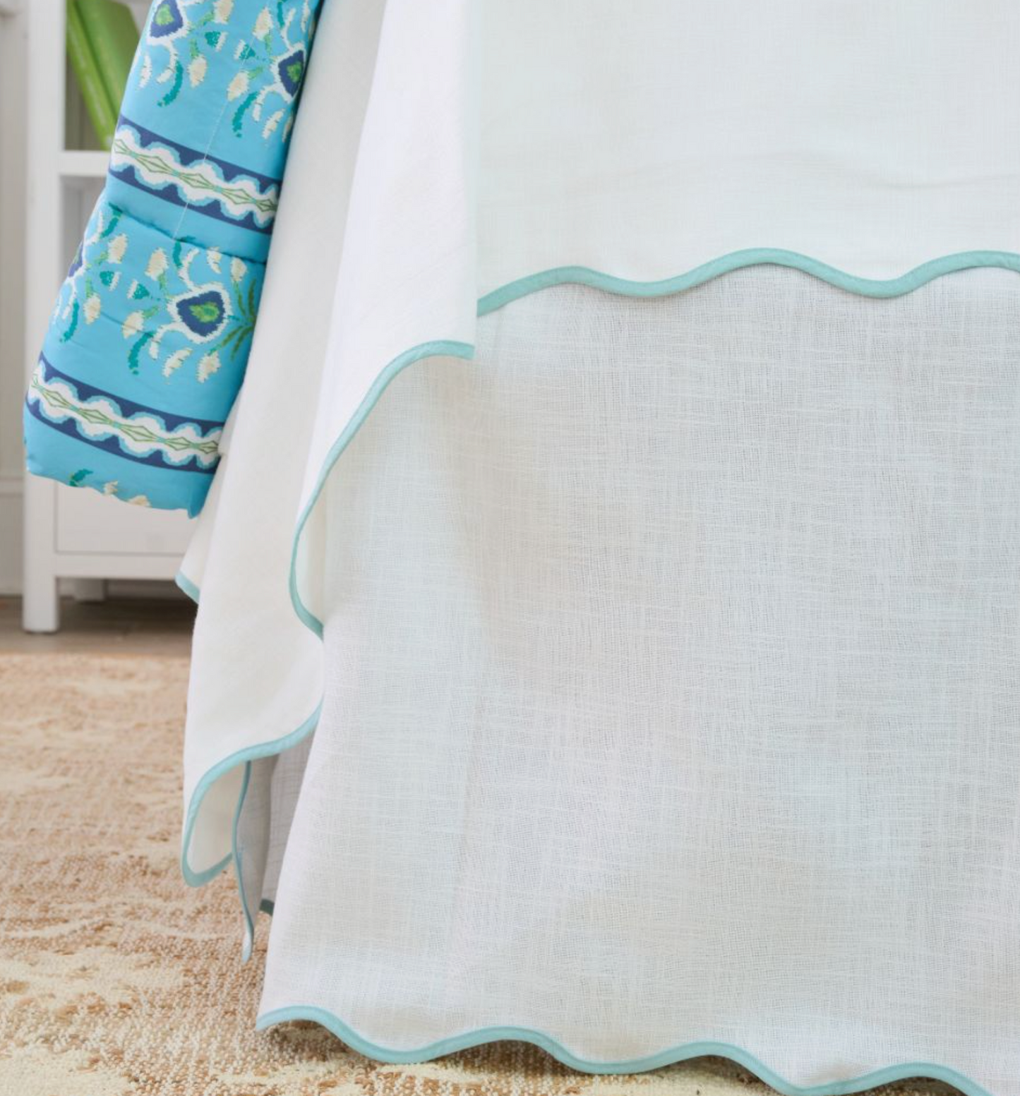A close-up of a white textured bed skirt with a scalloped light blue edge, shown next to a matching blue quilt with a floral and wave pattern.