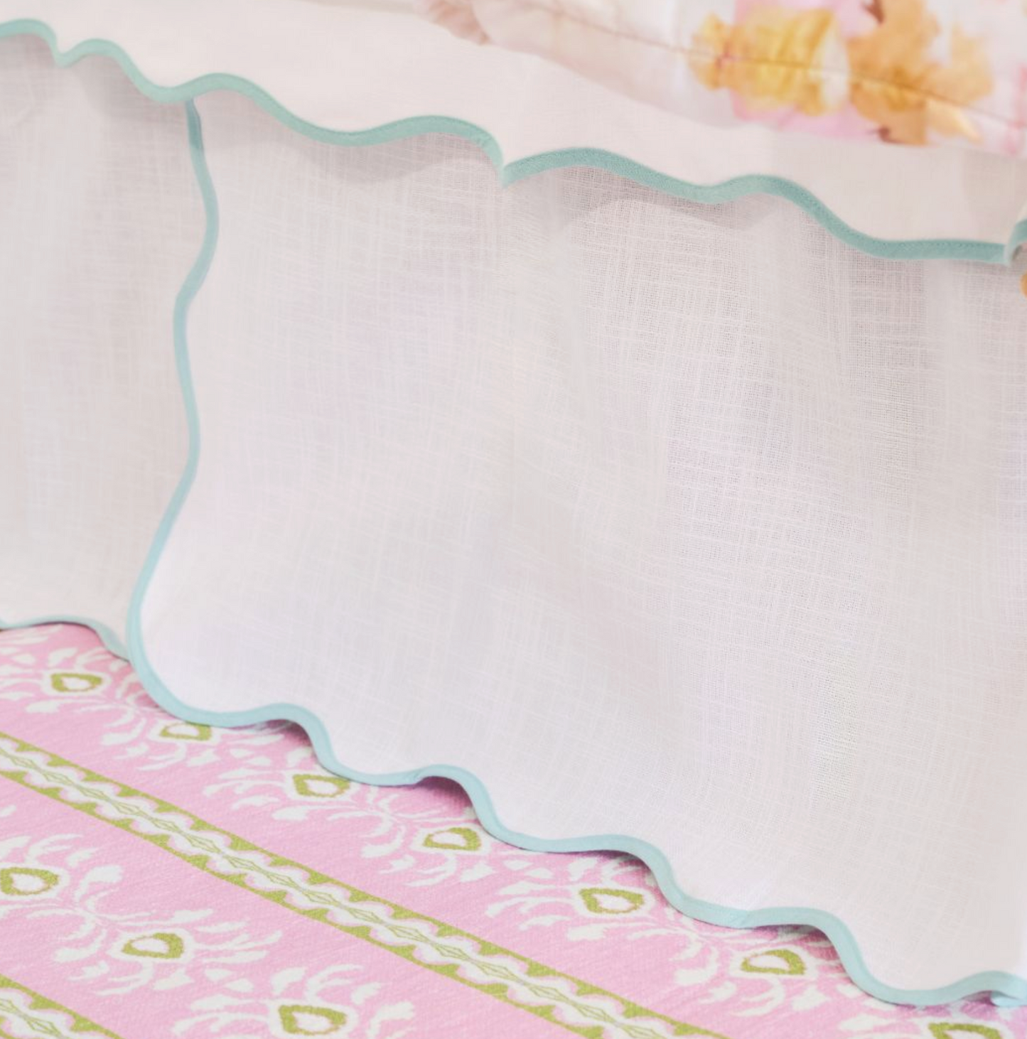 A close-up of a white textured fabric skirt or drape with a scalloped edge and light blue trim, layered over a pink patterned textile.