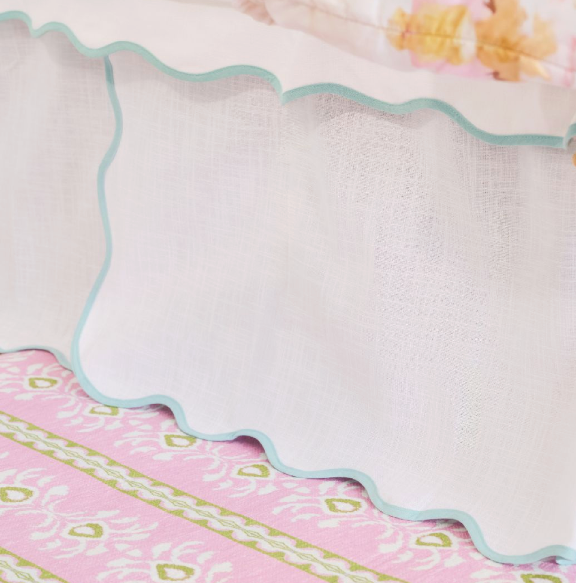A close-up of a white textured fabric skirt or drape with a scalloped edge and light blue trim, layered over a pink patterned textile.