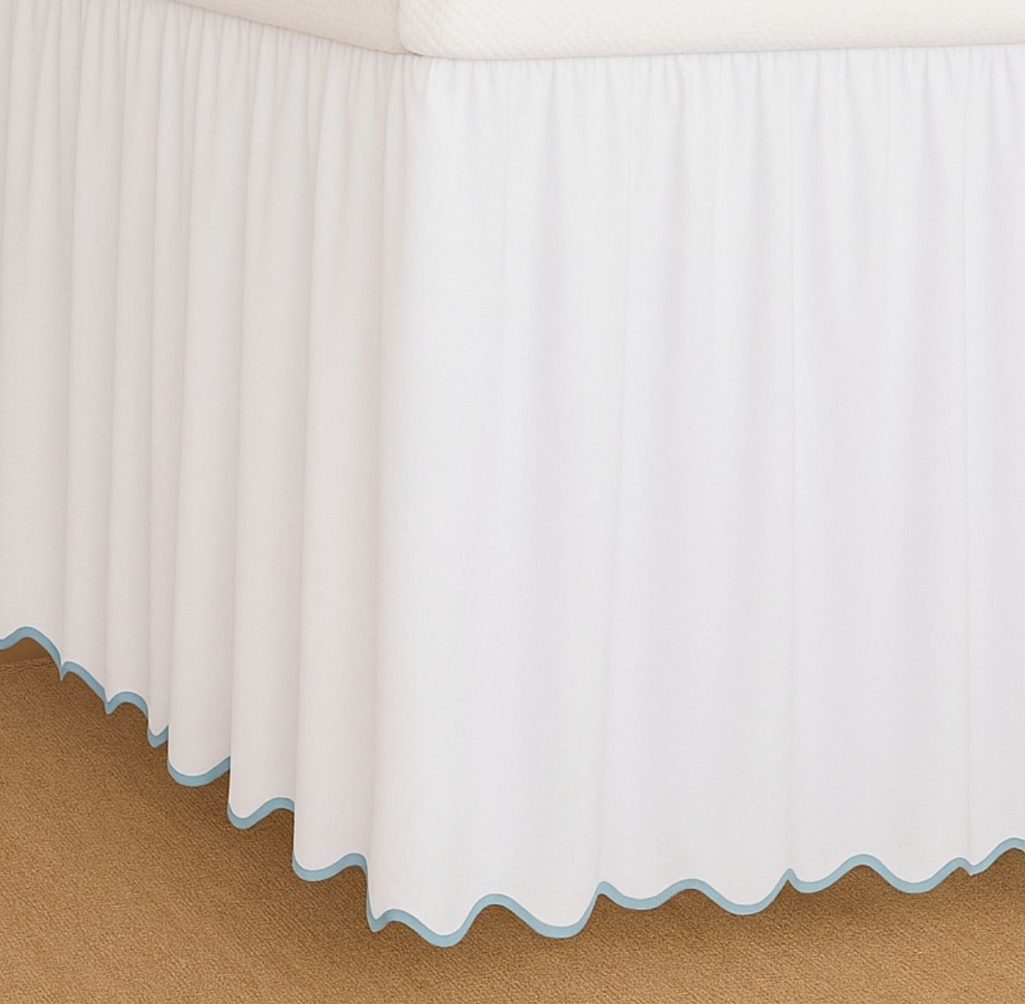 White bed skirt with a scalloped light blue edge trim, shown draped over the corner of a bed on a tan carpeted floor.