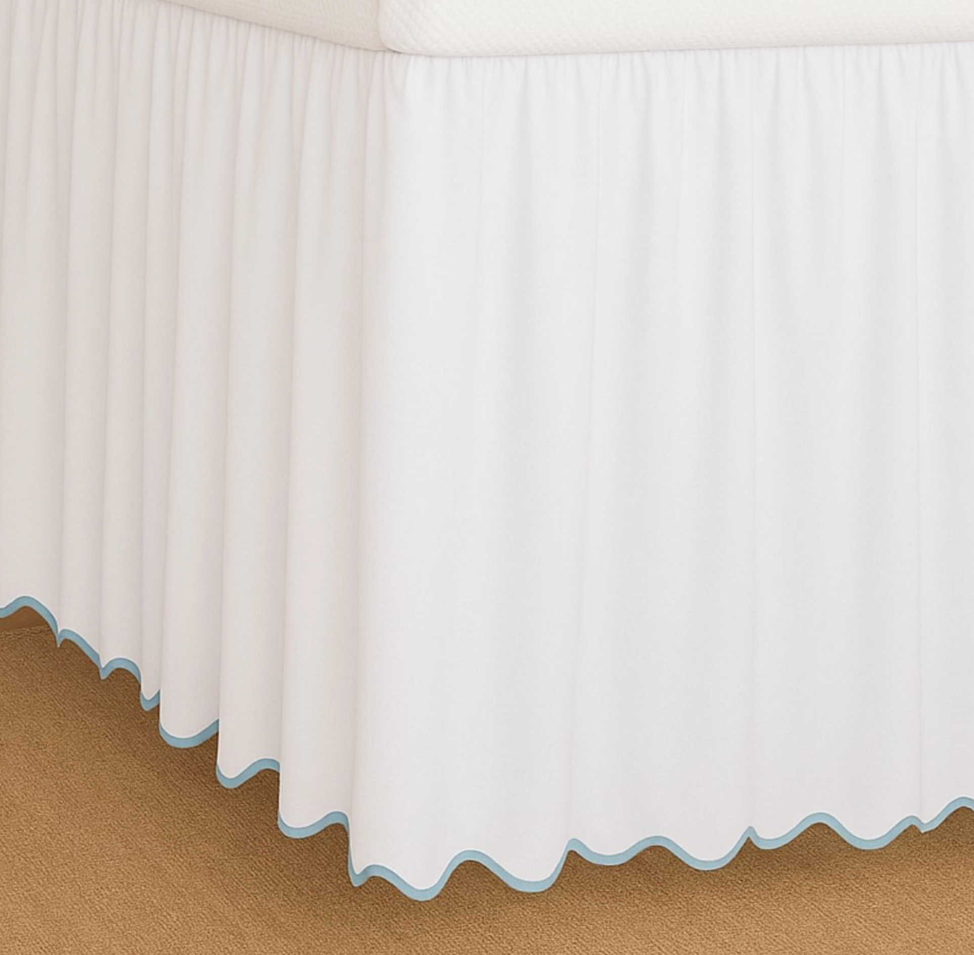 White bed skirt with a scalloped light blue edge trim, shown draped over the corner of a bed on a tan carpeted floor.