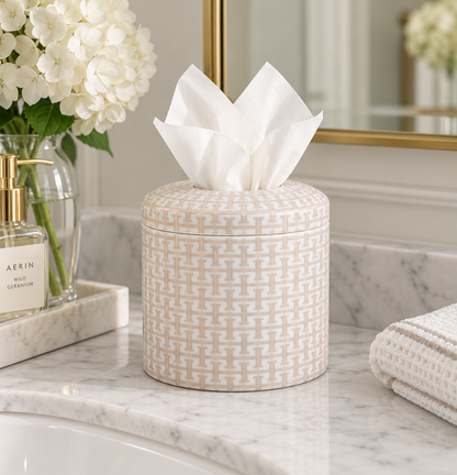 A round tissue box cover with a beige and white geometric monogram pattern, holding white tissue paper, set on a white marble bathroom counter next to a glass vase of white flowers and a folded white towel.