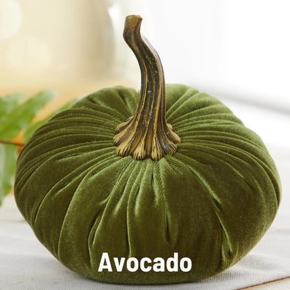 An avocado green velvet decorative pumpkin with a textured faux-stem and the word 'Avocado' printed at the bottom, set against a softly blurred indoor background.