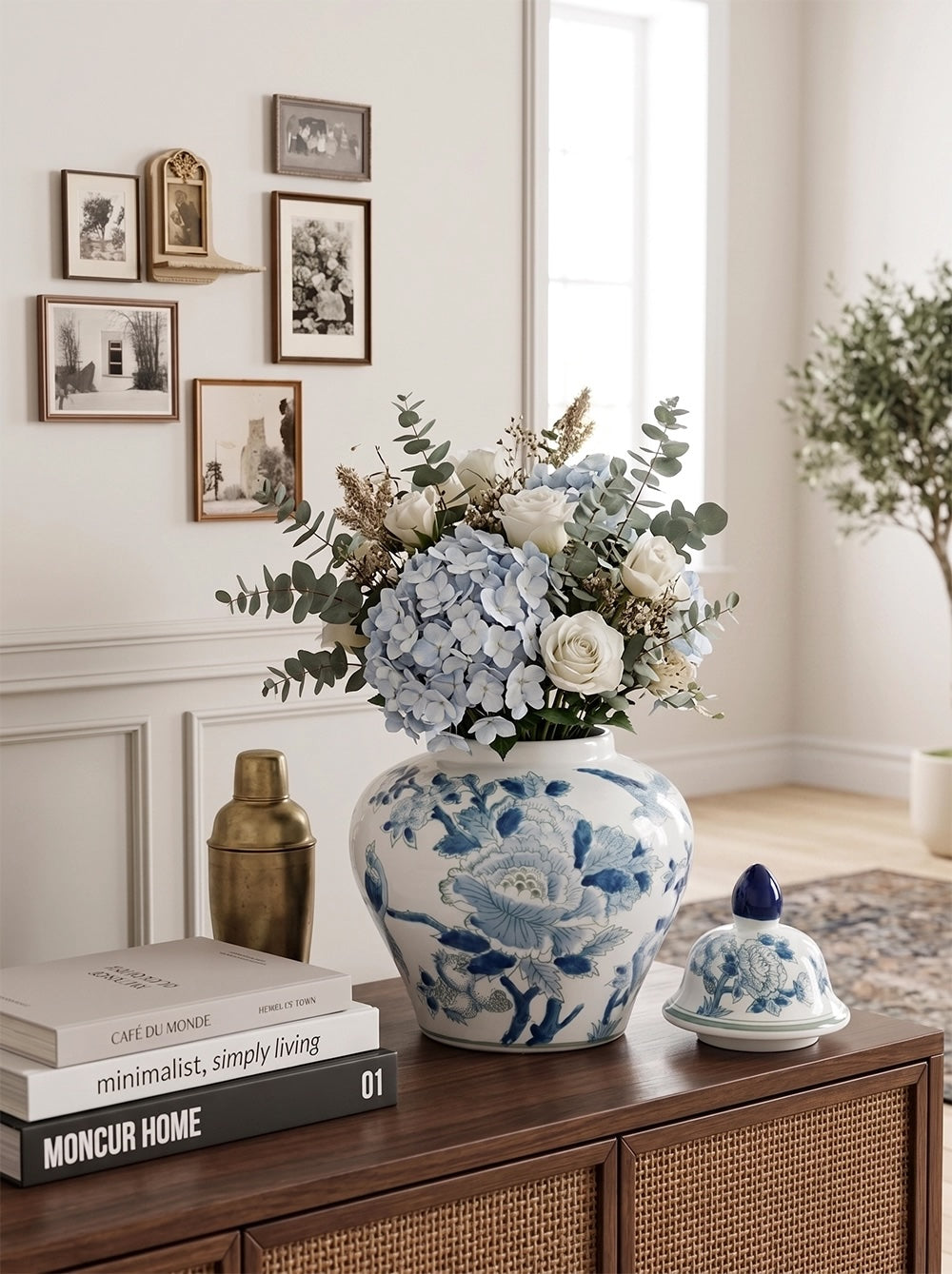 A blue and white floral ceramic vase filled with hydrangeas and greenery sits on a wooden console table in a bright, styled room. A matching blue and white lidded ceramic container and stacked books are nearby, with framed wall art and a plant in the background.