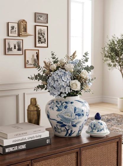 A blue and white floral ceramic vase filled with hydrangeas and greenery sits on a wooden console table in a bright, styled room. A matching blue and white lidded ceramic container and stacked books are nearby, with framed wall art and a plant in the background.