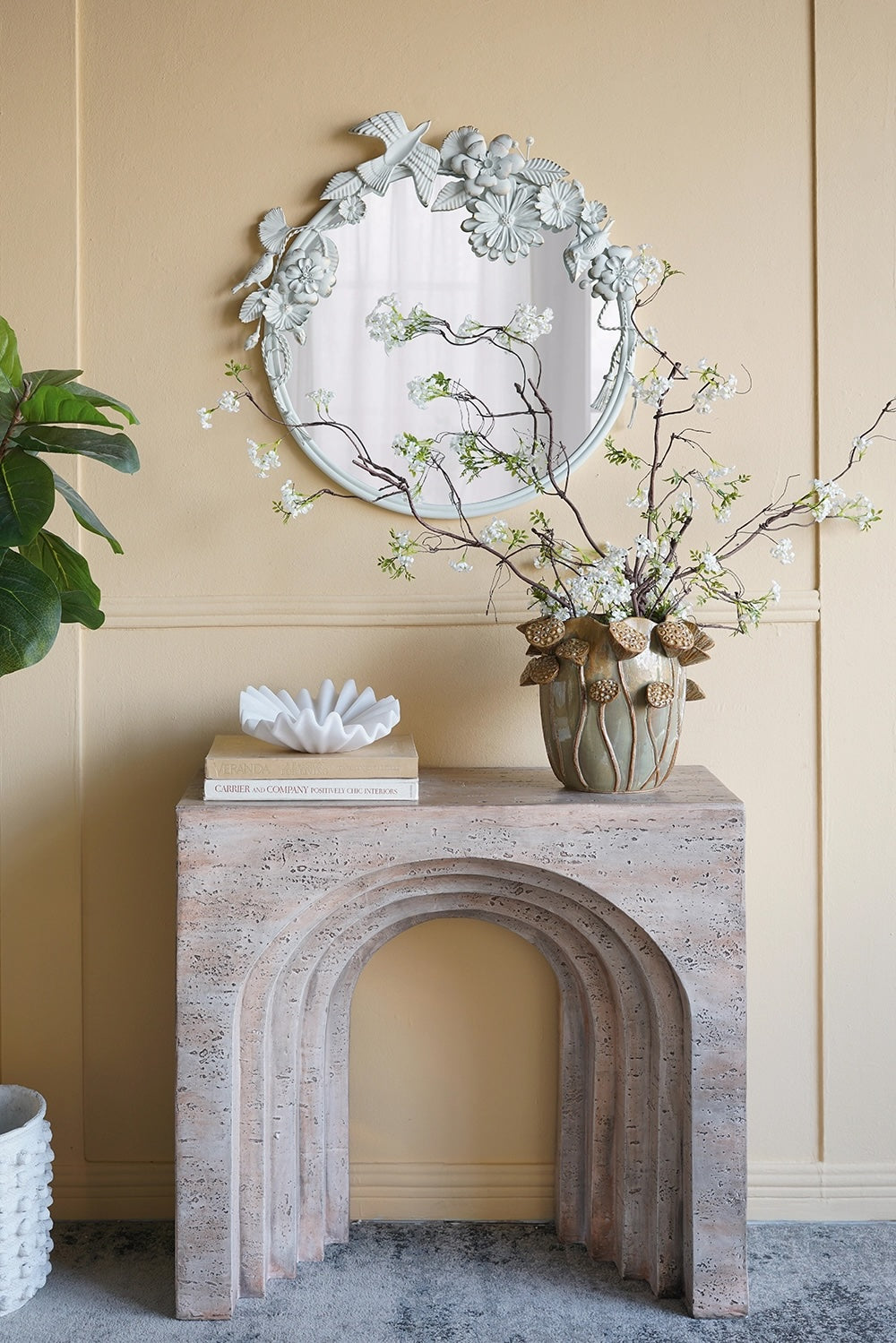 Decorative mirror with floral design above a stone fireplace mantel with a vase of flowers and books.