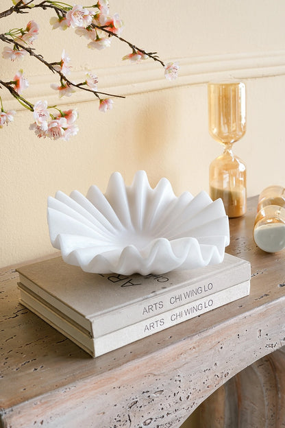 A white ceramic shell-shaped bowl with a fluted, scalloped edge sits on top of two stacked books on a rustic wooden table. In the background, a gold hourglass and a small white candle are visible against a warm beige wall with cherry blossom branches in the upper left corner.