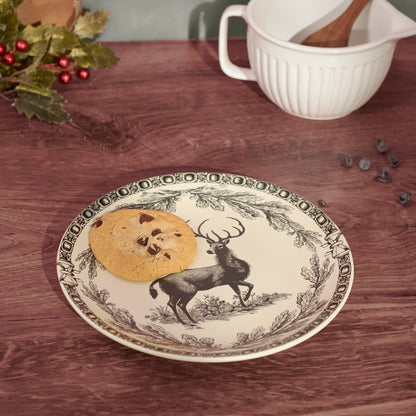 A round ceramic plate with a black and white deer and foliage border design, shown on a wooden table with a cookie on it. A white ribbed mug with a wooden spoon sits in the background, along with some holiday greenery and blueberries.