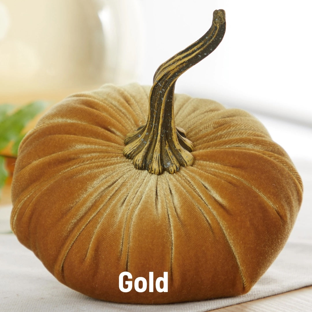 A decorative gold velvet pumpkin with a dark textured stem, resting on a light fabric surface with a soft blurred background. The word 'Gold' is printed in white text at the bottom.