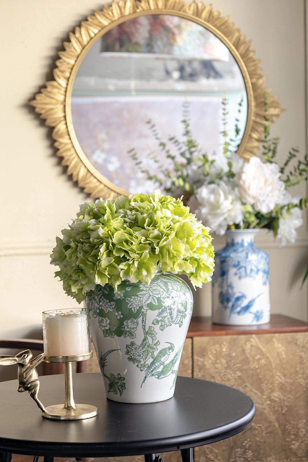 A green hydrangea arrangement in a white ceramic vase with a green botanical pattern, displayed on a black side table in a styled interior setting with a gold mirror and blue-and-white porcelain vase in the background.