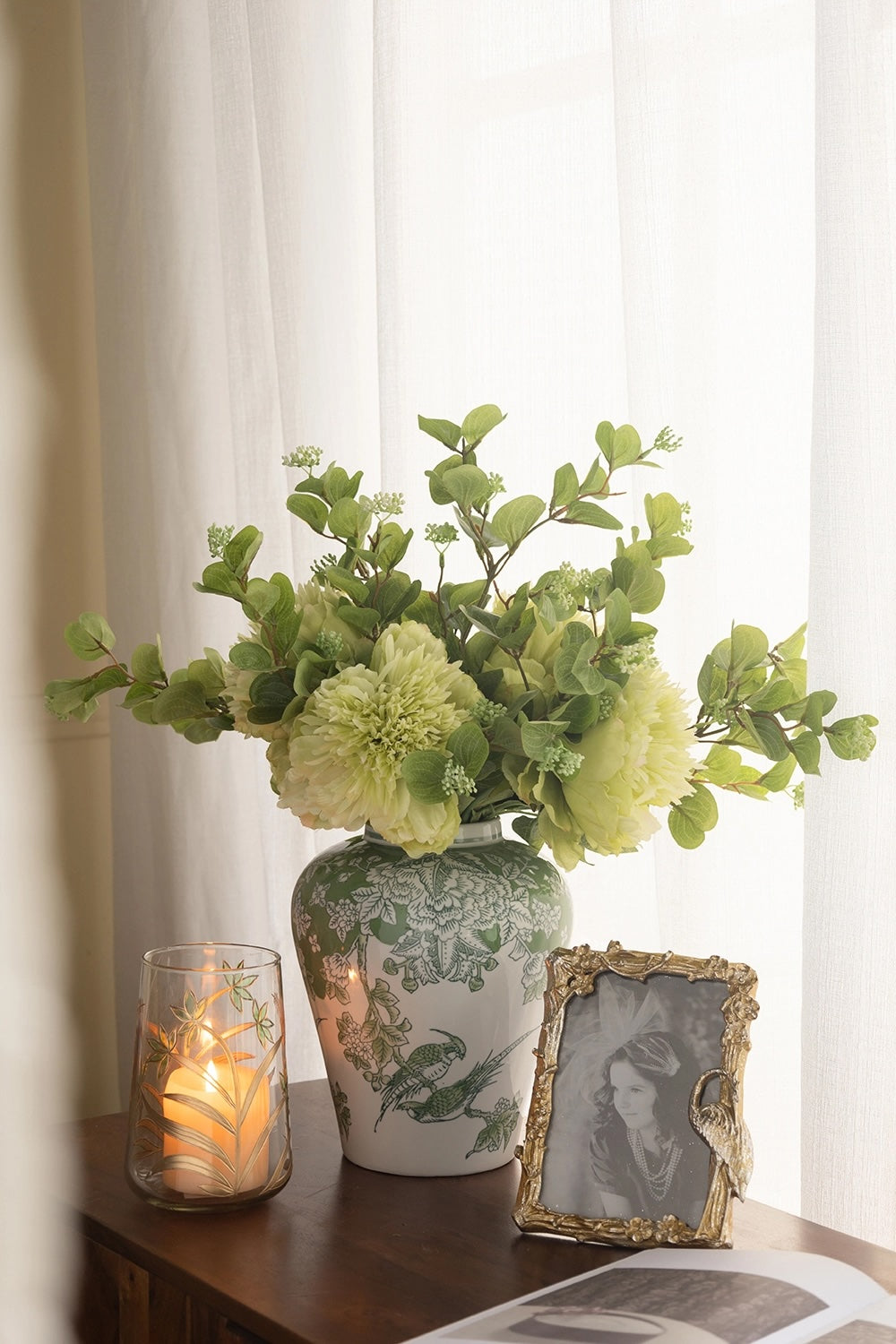 A green floral arrangement in a white ceramic vase with green botanical patterns, placed on a wooden table beside a lit candle in a glass holder and a small ornate gold picture frame against a soft white curtain background.