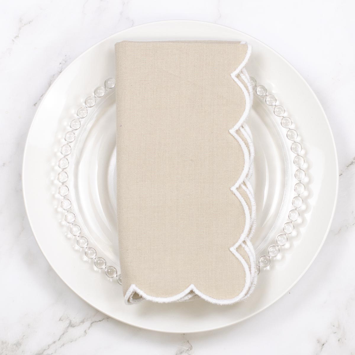 A folded beige linen napkin with a white scalloped edge, displayed on a white plate with a clear beaded charger plate on a white marble surface.