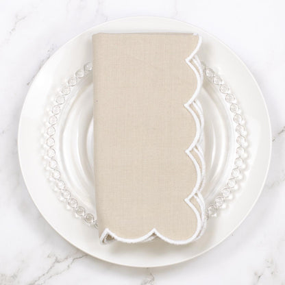 A folded beige linen napkin with a white scalloped edge, displayed on a white plate with a clear beaded charger plate on a white marble surface.