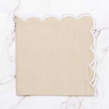 A square beige napkin with a scalloped white edge, laid flat on a white marble surface.