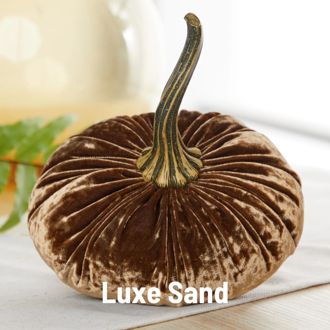 A plush velvet decorative pumpkin in a warm brown shade labeled 'Luxe Sand' at the bottom, featuring a ribbed surface and a textured stem, displayed on a light surface with a soft background.