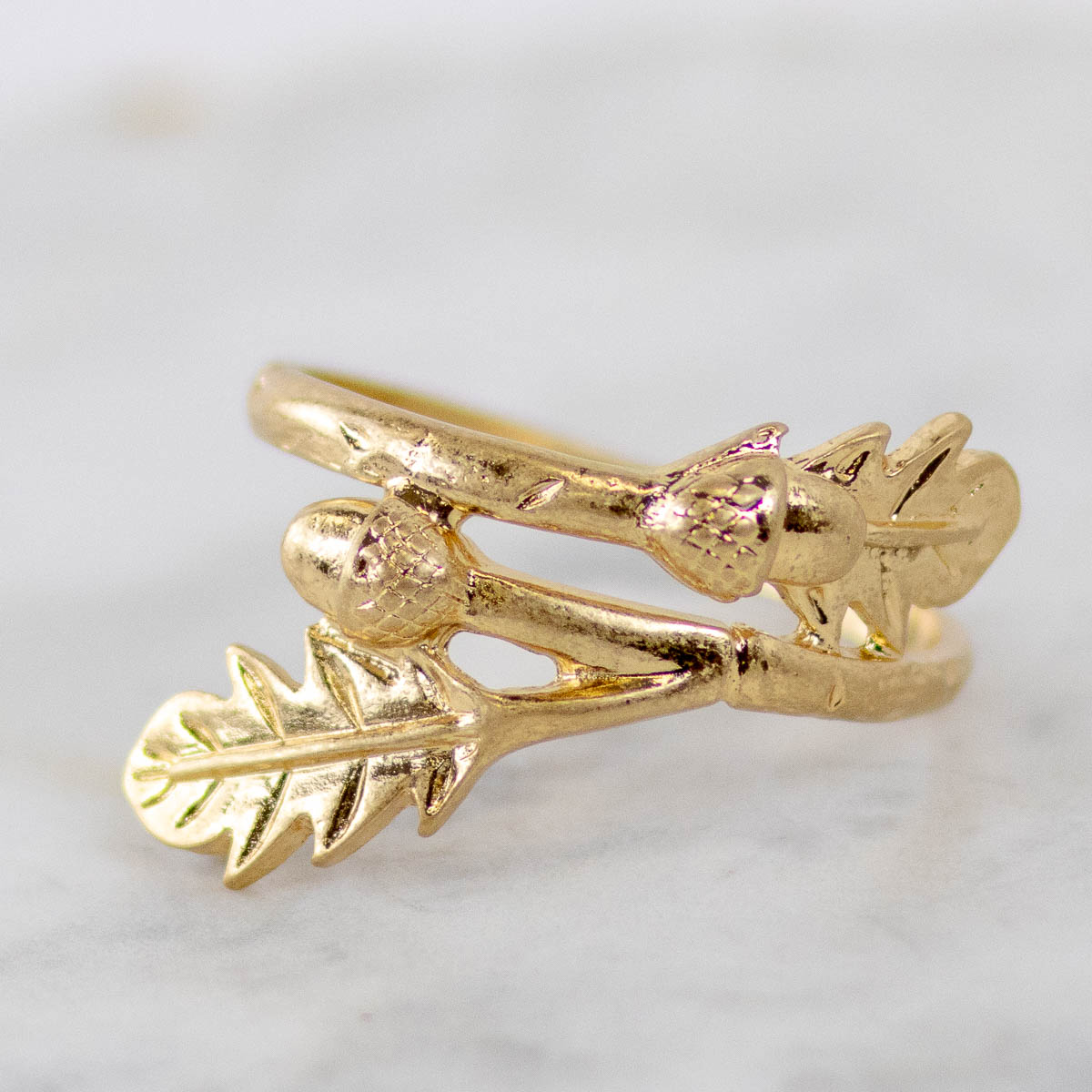 A gold ring designed to look like a branch with acorns and leaves, resting on a light gray marble surface.