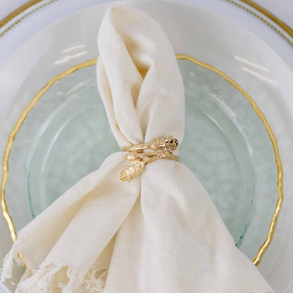 A cream-colored linen napkin tied with a gold leaf-shaped ring, resting on a clear glass plate with a gold-trimmed white ceramic plate.