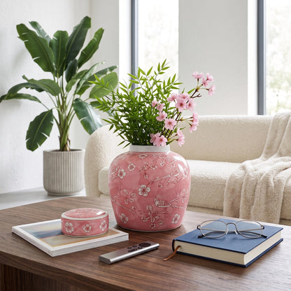 A pink ceramic vase with a white floral pattern sits on a wooden coffee table in a bright living room, filled with green foliage and pink flowers. A matching pink container, a remote, a blue book, and glasses are on the table, with a cream sofa and a potted plant in the background.