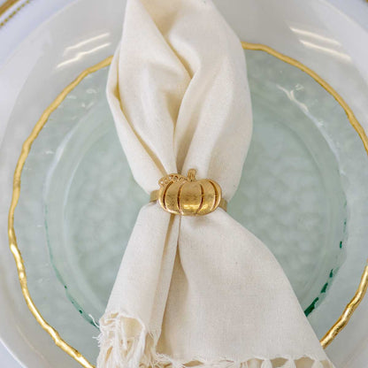 An off-white fabric napkin tied with a small metallic gold pumpkin ring, resting on a light aqua textured plate with a gold-trimmed white charger plate.