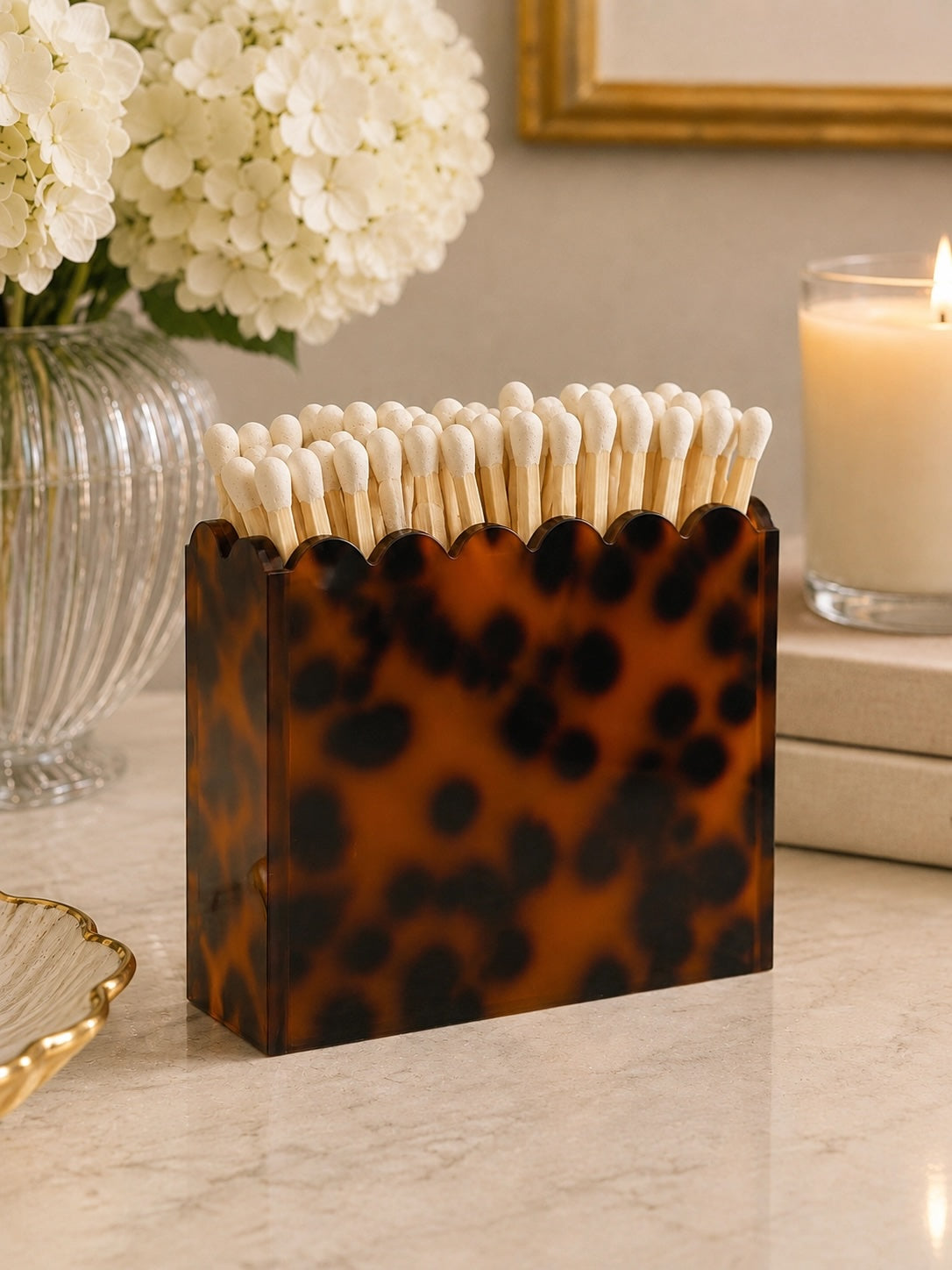 A tortoiseshell-patterned matchbox holder filled with white cotton matches, styled on a marble tabletop with a glass vase of white flowers and a lit candle in the background.