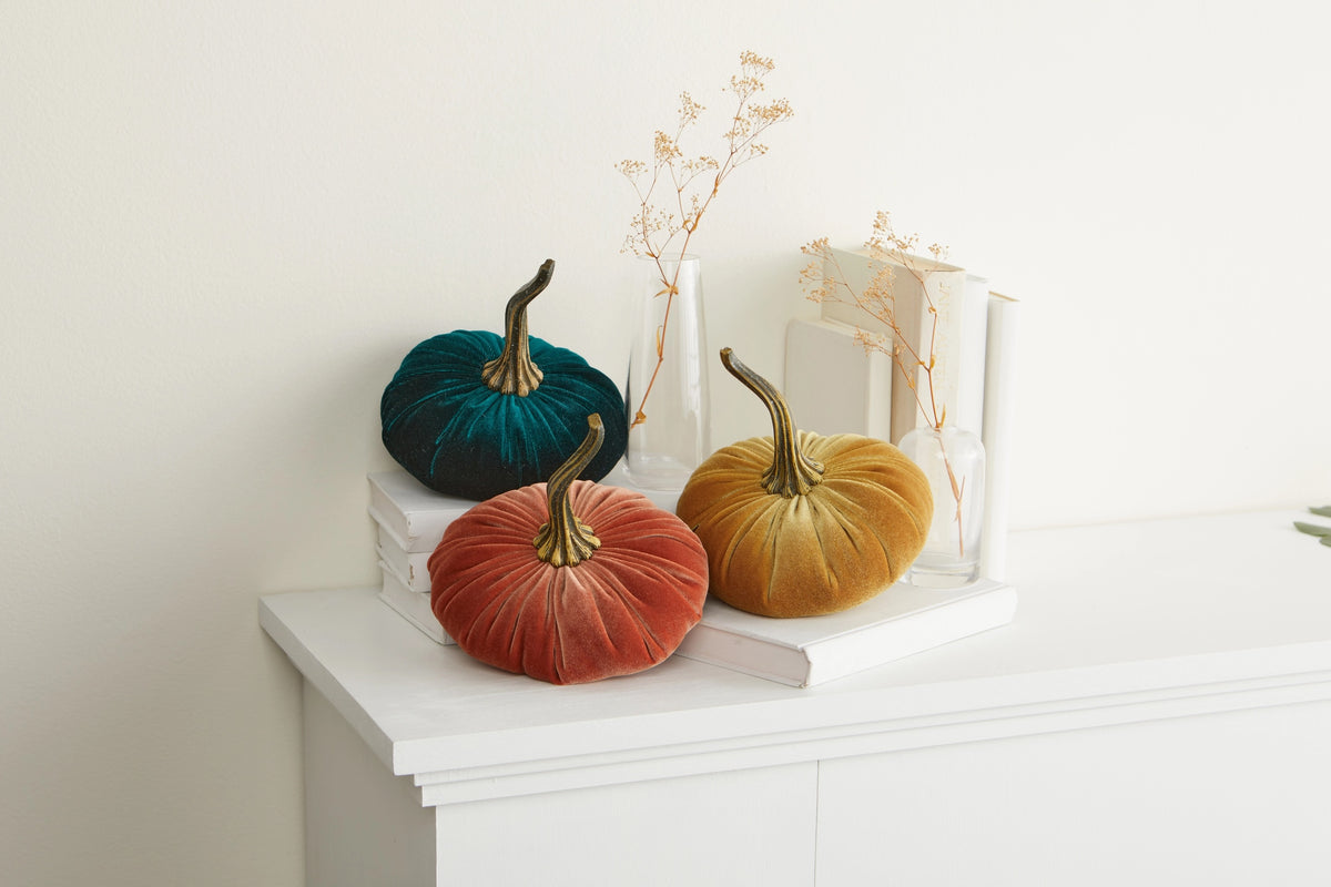 Three velvet decorative pumpkins in teal, rust orange, and mustard yellow with brass-colored stems, styled on white books and a white console table against a warm off-white wall.