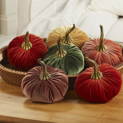 Six velvet decorative pumpkins in assorted fall colors, including red, green, dusty rose, mustard, and terracotta, arranged in and around a woven basket on a wooden table with a soft white sofa in the background.